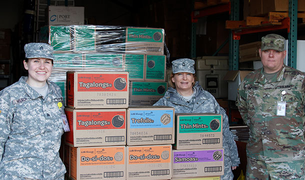 Three US Army Members stand in front of plastic wrapped cases of Girl Scout Cookies