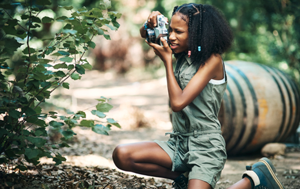Girl kneeling in the woods to take a picture of a bush.