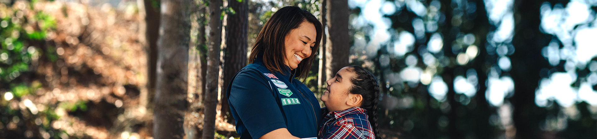  adult volunteer in vest outside hugging girl scout while hiking 